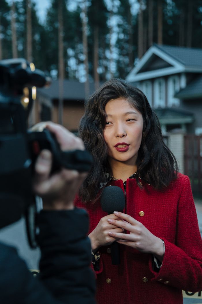 Services Asian woman reporting news on camera outdoors, wearing red coat.