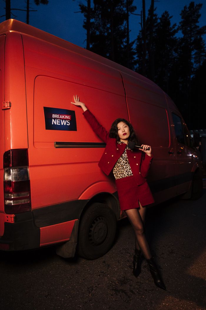 Services Asian woman in red attire posing confidently by an orange news van at night.